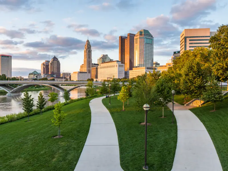 Photo of downtown Columbus, Ohio with 2 paths running along the river and tall buildings in the background.
